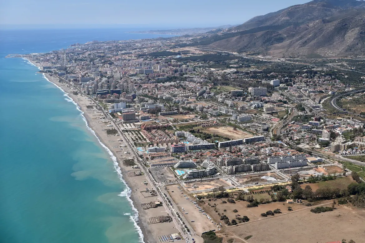 Eine Luftaufnahme der Küste von Torremolinos, ein langer Strand mit der Stadt dahinter und einem in der Ferne aufragenden Gebirgskamm