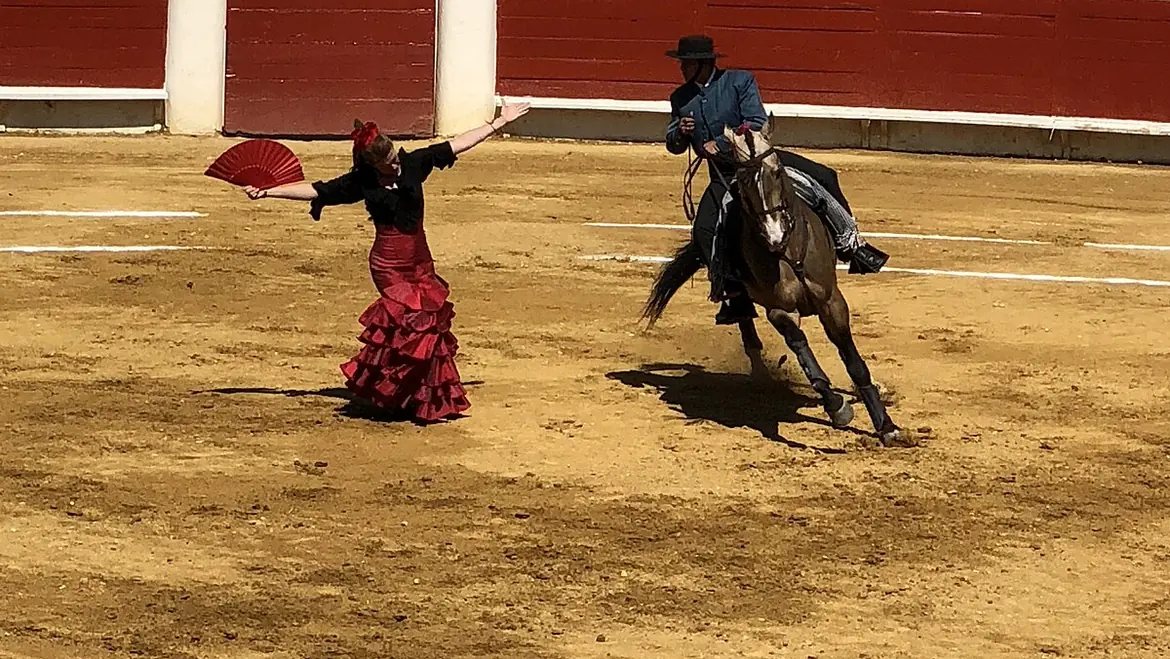 Eine Flamenco-Tänzerin in einem roten Rüschenrock hält einen roten Fächer auf dem Sand einer Arena, während ein Reiter auf einem dunklen Pferd neben ihr kreist