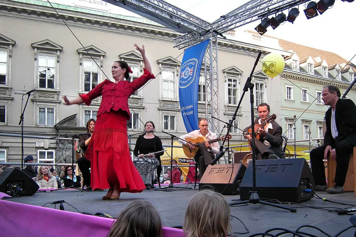 Auf einer Festivalbühne posiert eine Tänzerin in einem roten Kleid, hinter ihr sitzen zwei Gitarristen und ein Sänger