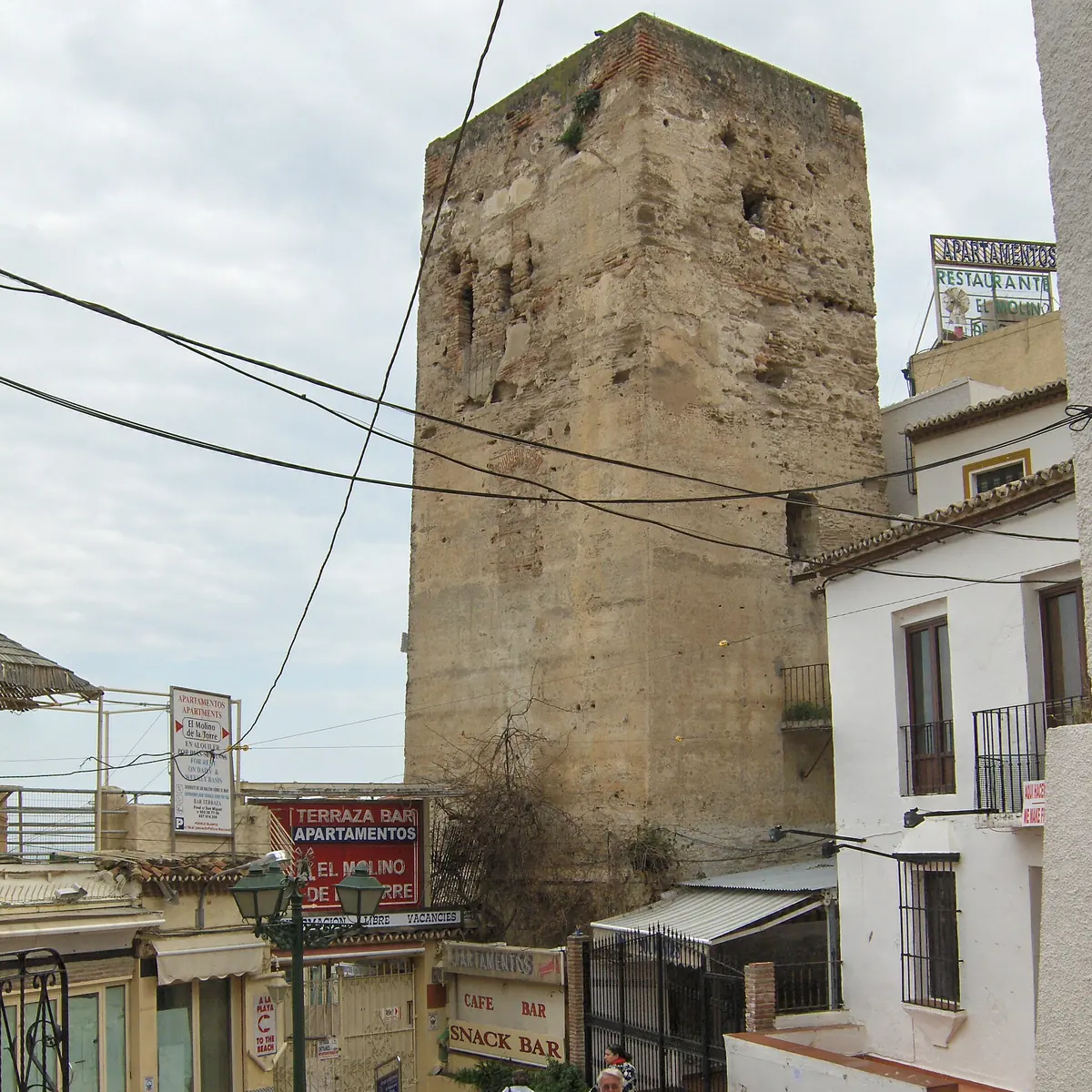 Der viereckige Steinturm in Torremolinos, der sich über weiß getünchte Häuser und eine schmale Gasse mit Ladenschildern erhebt